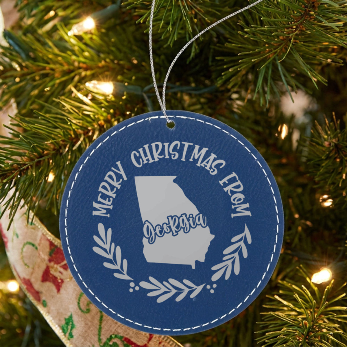 Blue leatherette round Christmas ornament engraved Merry Christmas From Georgia, with the state shape in a laurel wreath, hanging on a tree