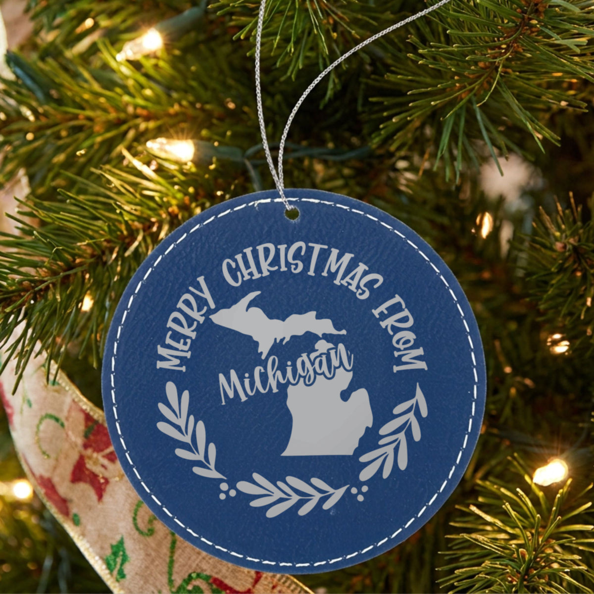 Blue leatherette round Christmas ornament engraved Merry Christmas From Michigan, with the state shape in a laurel wreath, hanging on a tree