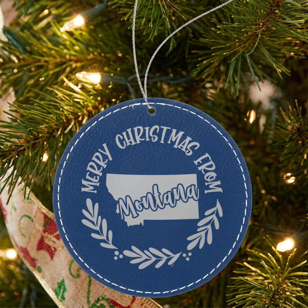 Blue leatherette round Christmas ornament engraved Merry Christmas From Montana, with the state shape in a laurel wreath, hanging on a tree