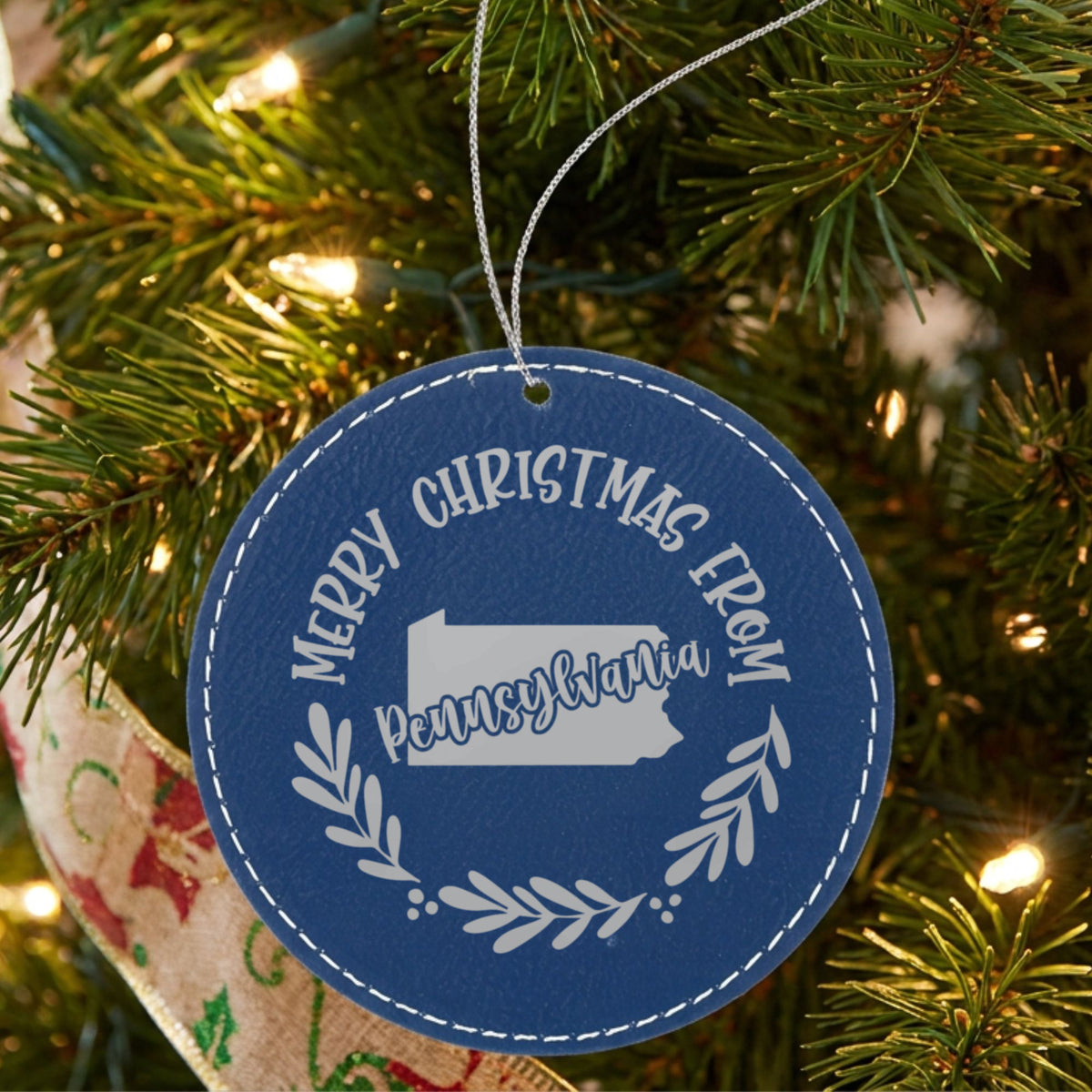 Blue leatherette round Christmas ornament engraved Merry Christmas From Pennsylvania, with the state shape in a laurel wreath, hanging on a tree