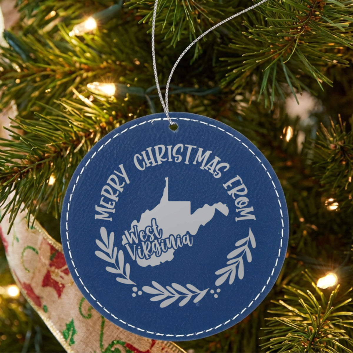 Blue leatherette round Christmas ornament engraved Merry Christmas From West Virginia, with the state shape in a laurel wreath, hanging on a tree