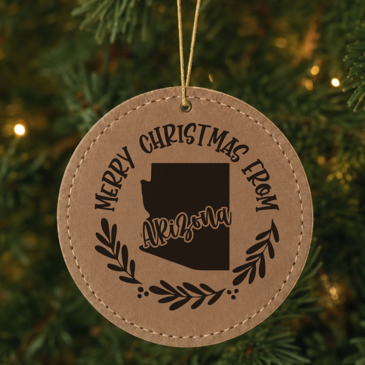 Brown leatherette round Christmas ornament engraved Merry Christmas From Arizona, with the state shape in a laurel wreath, hanging on a tree