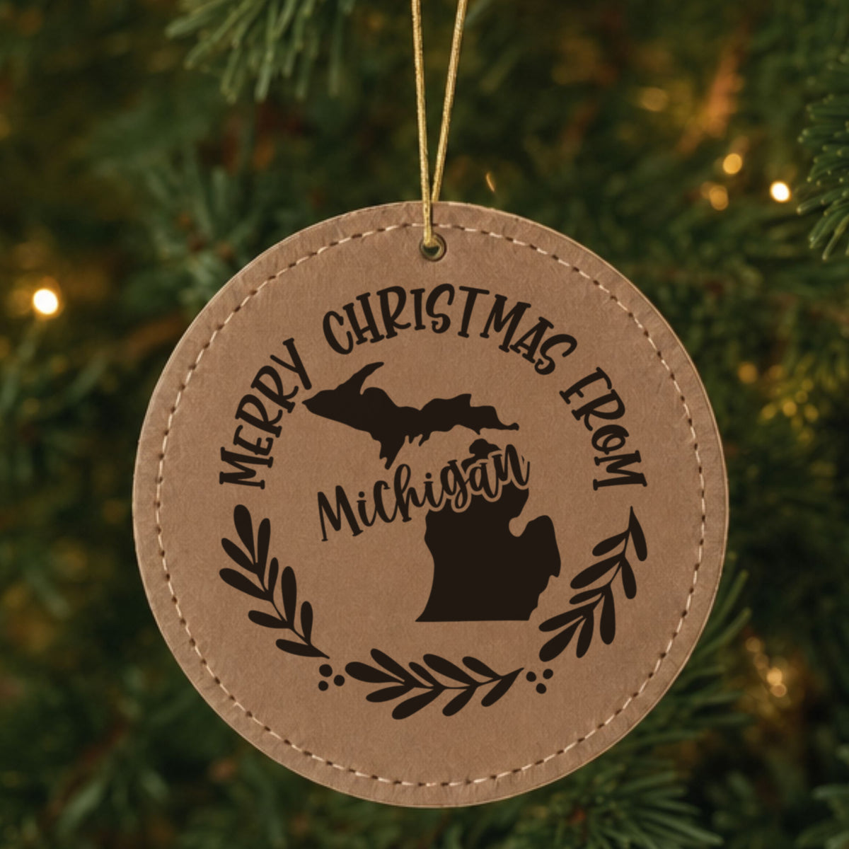 Brown leatherette round Christmas ornament engraved Merry Christmas From Michigan, with the state shape in a laurel wreath, hanging on a tree