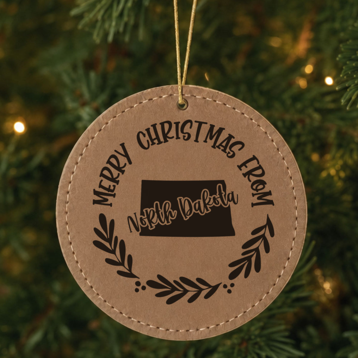 Brown leatherette round Christmas ornament engraved Merry Christmas From North Dakota, with the state shape in a laurel wreath, hanging on a tree