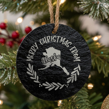 Round slate Christmas ornament engraved Merry Christmas From Alaska, with the state shape in a laurel wreath