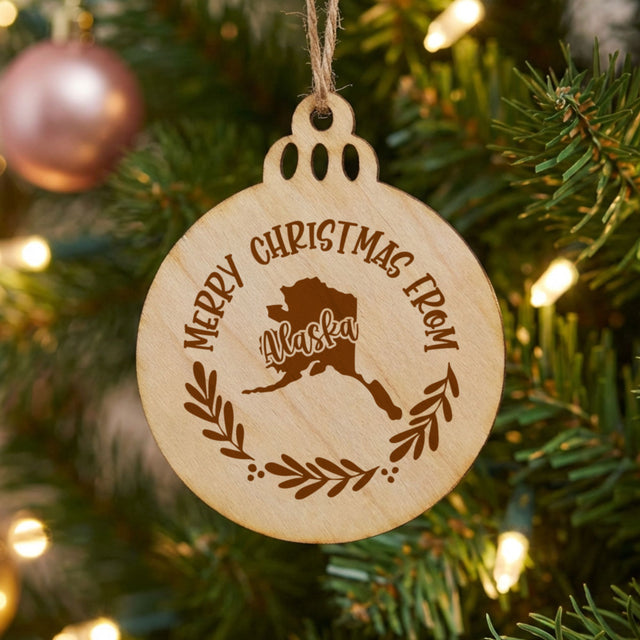 Round wood Christmas ornament engraved Merry Christmas From Alaska, with the state shape in a laurel wreath