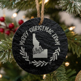 Round slate Christmas ornament engraved Merry Christmas From Idaho, with the state shape in a laurel wreath