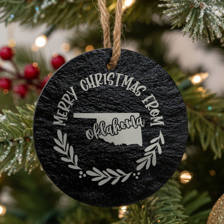 Round slate Christmas ornament engraved Merry Christmas From Oklahoma, with the state shape in a laurel wreath
