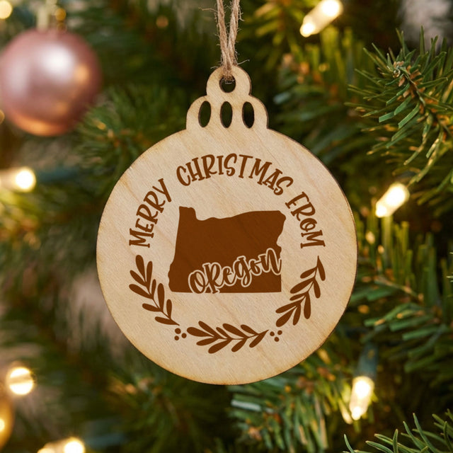 Round wood Christmas ornament engraved Merry Christmas From Oregon, with the state shape in a laurel wreath