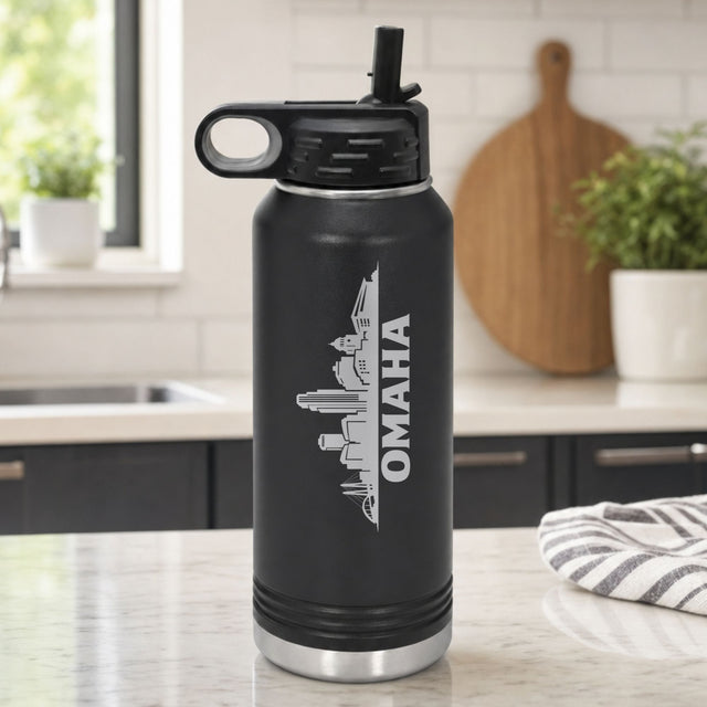 Black 32 oz insulated water bottle laser engraved with the Omaha, Nebraska skyline running up the side, standing on a kitchen counter