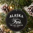 Black leatherette round Christmas ornament laser engraved with an Alaska design of a grizzly bear crossing the state map, hanging on a tree