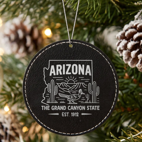 Black leatherette round Christmas ornament laser engraved with an Arizona design of saguaro cactus, a roadrunner and the Grand Canyon at sunrise, hanging on a tree