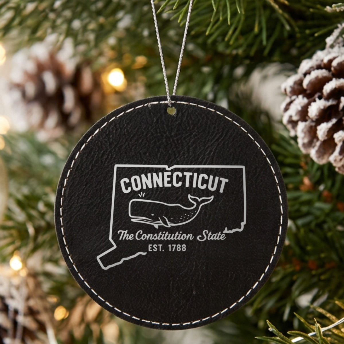 Black leatherette round Christmas ornament laser engraved with a Connecticut design of a sperm whale, hanging on a tree