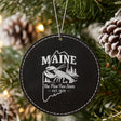 Black leatherette round Christmas ornament laser engraved with a Maine design of a lobster, pines and a lighthouse, hanging on a tree