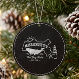 Black leatherette round Christmas ornament laser engraved with a Massachusetts design of a whale, a lighthouse and the sun over Cape Cod and the islands, hanging on a tree