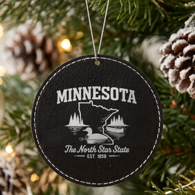 Black leatherette round Christmas ornament laser engraved with a Minnesota design of a loon, a canoe and lakeside pines, hanging on a tree