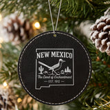Black leatherette round Christmas ornament laser engraved with a New Mexico design of a roadrunner, cactus and pines, hanging on a tree