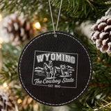 Black leatherette round Christmas ornament laser engraved with a Wyoming design of a cowboy on horseback, a bighorn sheep and mountains, hanging on a tree