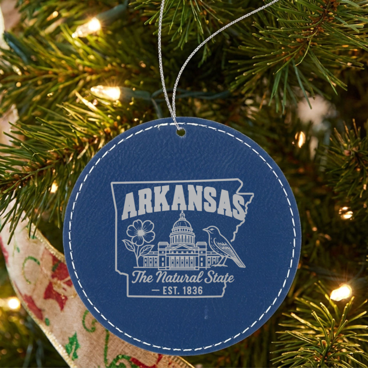 Blue leatherette round Christmas ornament laser engraved with an Arkansas design of the capitol dome, an apple blossom and a mockingbird, hanging on a tree