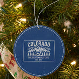 Blue leatherette round Christmas ornament laser engraved with a Colorado design of snowcapped peaks, pine forest and a bighorn sheep, hanging on a tree