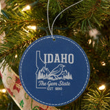 Blue leatherette round Christmas ornament laser engraved with an Idaho design of a mountain bluebird, mountains and pines, hanging on a tree