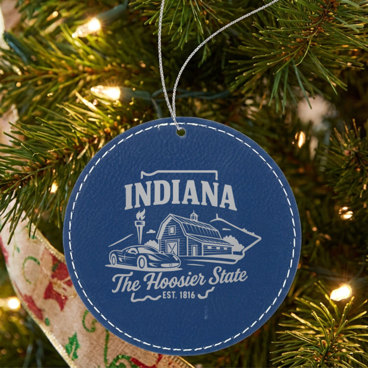 Blue leatherette round Christmas ornament laser engraved with an Indiana design of a red barn, a race car and a torch, hanging on a tree
