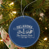 Blue leatherette round Christmas ornament laser engraved with an Oklahoma design of a cowboy hat, an oil derrick and a bison, hanging on a tree