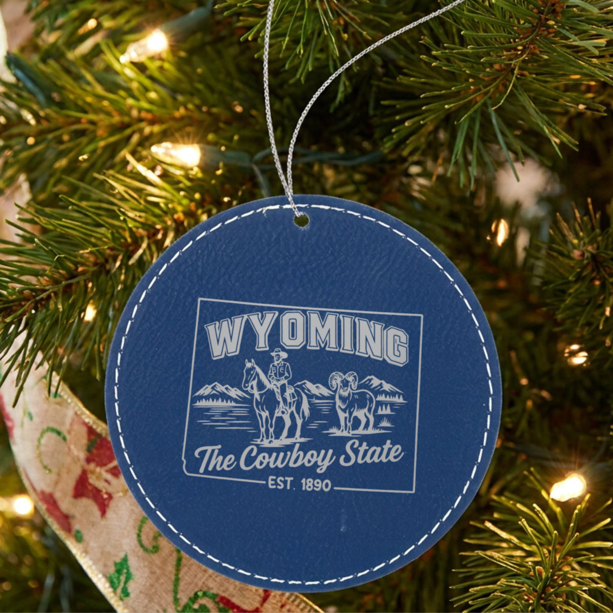 Blue leatherette round Christmas ornament laser engraved with a Wyoming design of a cowboy on horseback, a bighorn sheep and mountains, hanging on a tree