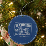 Blue leatherette round Christmas ornament laser engraved with a Wyoming design of a cowboy on horseback, a bighorn sheep and mountains, hanging on a tree