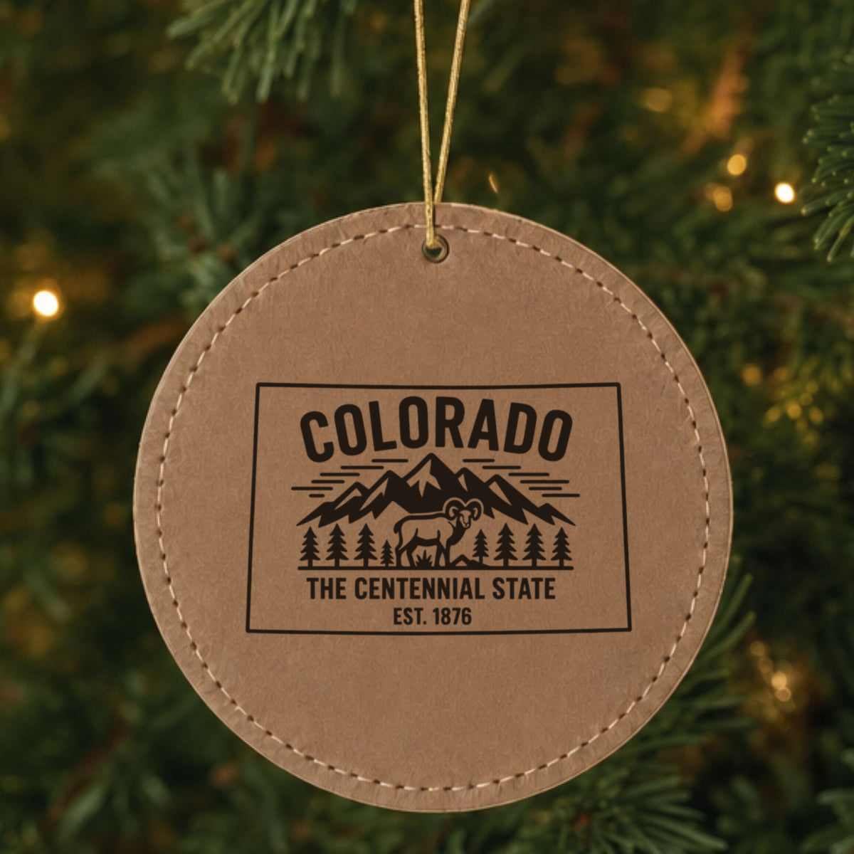 Brown leatherette round Christmas ornament laser engraved with a Colorado design of snowcapped peaks, pine forest and a bighorn sheep, hanging on a tree