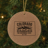 Brown leatherette round Christmas ornament laser engraved with a Colorado design of snowcapped peaks, pine forest and a bighorn sheep, hanging on a tree