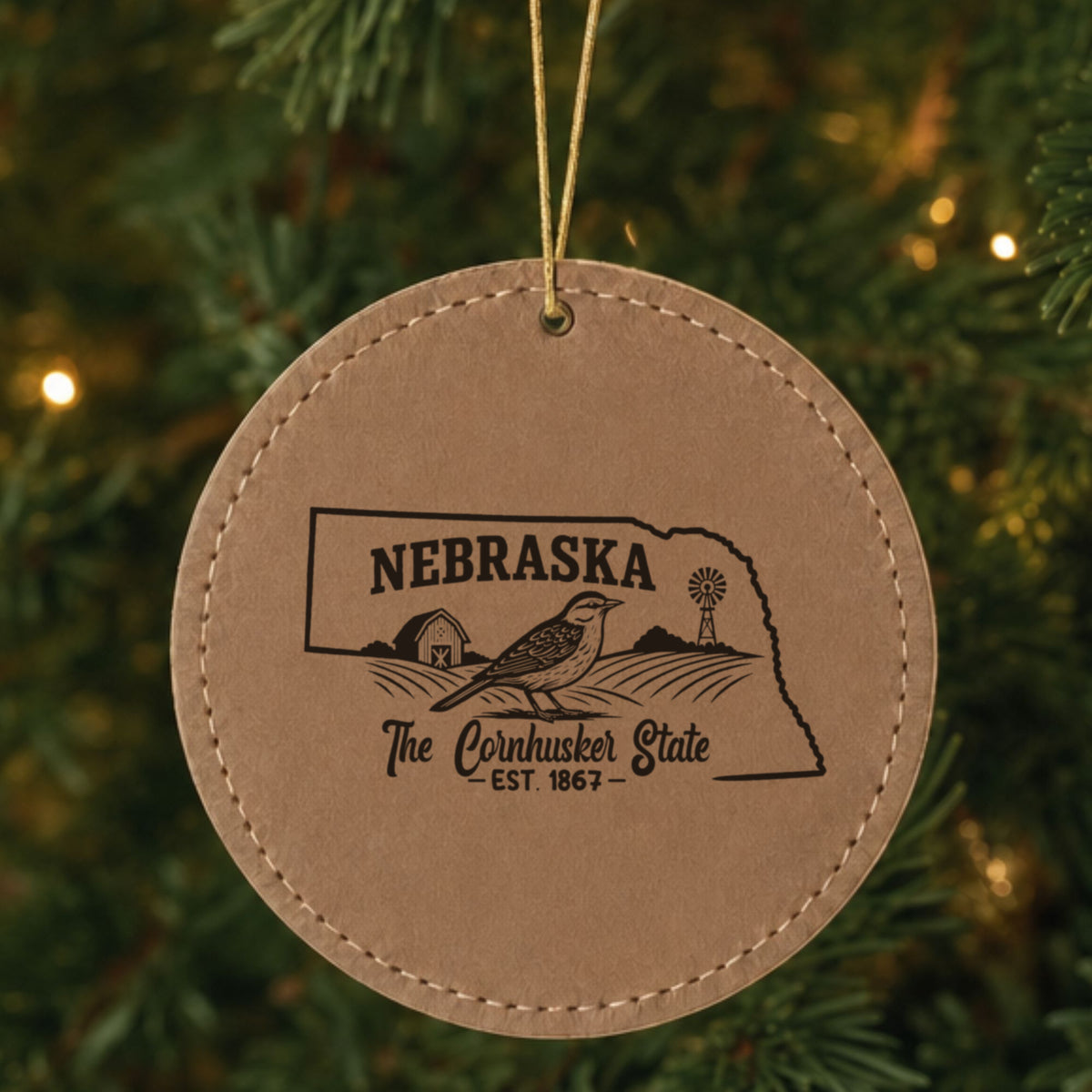 Brown leatherette round Christmas ornament laser engraved with a Nebraska design of a western meadowlark, a barn and a windmill, hanging on a tree