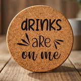 Round cork coaster laser engraved with "Drinks Are On Me", hand lettered design