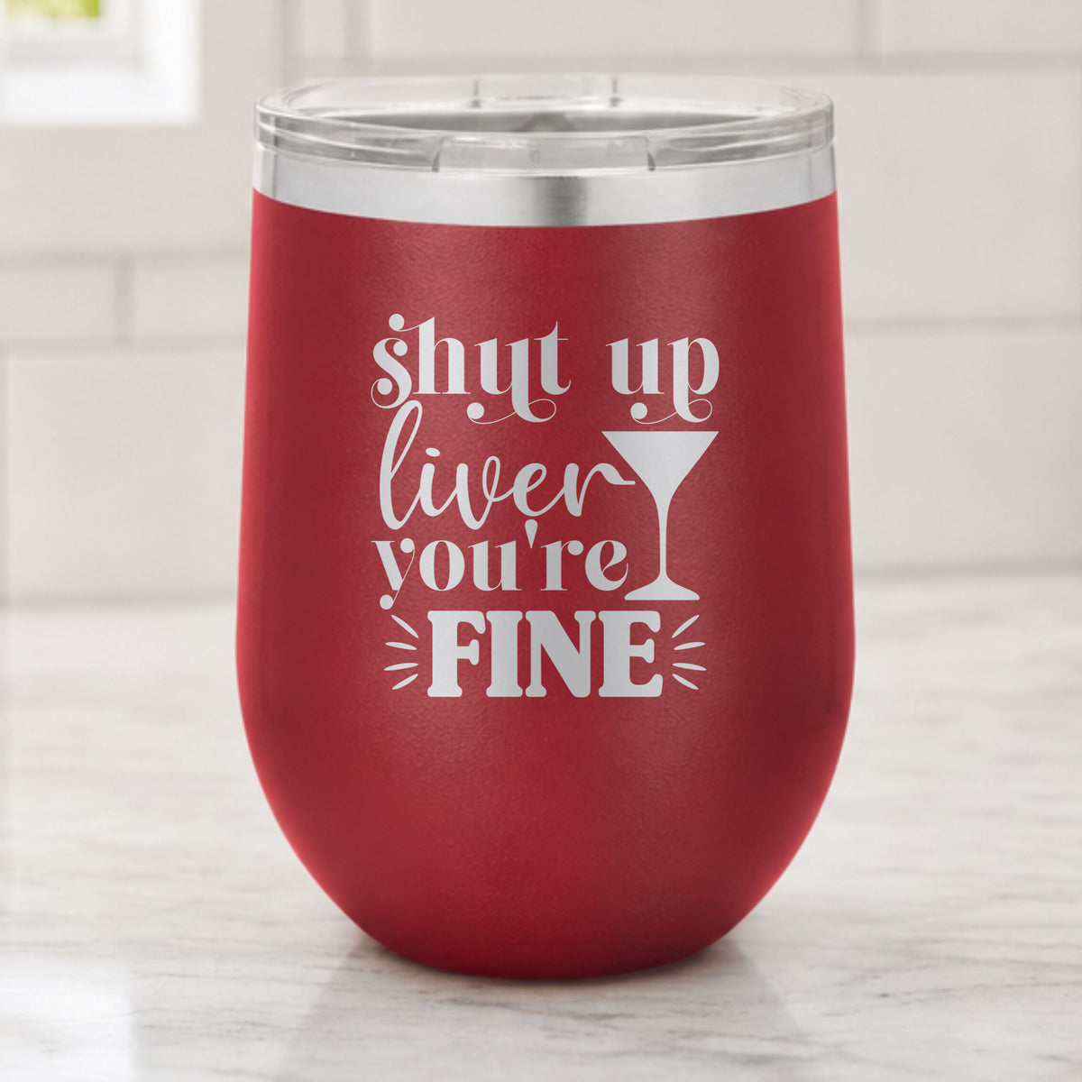 Maroon 12 oz stemless wine tumbler laser engraved with "Shut Up Liver, You're Fine", on a kitchen counter