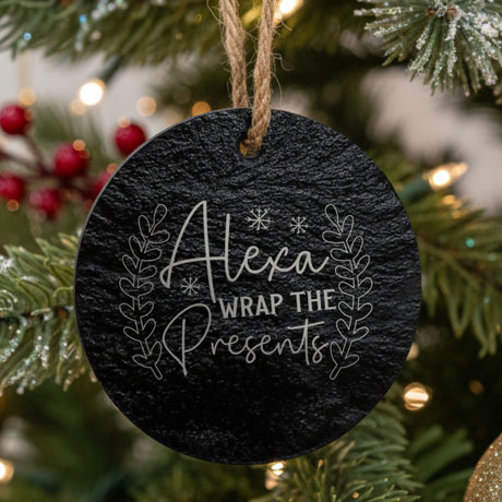 A round black slate Christmas ornament hanging from a jute string on a decorated Christmas tree. The ornament features white engraved text reading 'Alexa Wrap The Presents' surrounded by delicate leaf line drawings.