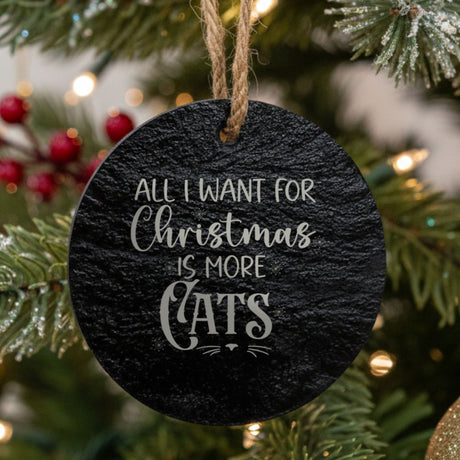 A round black slate Christmas ornament hanging from a jute string on a decorated Christmas tree. The ornament features the text 'ALL I WANT FOR Christmas IS MORE CATS' in white lettering.