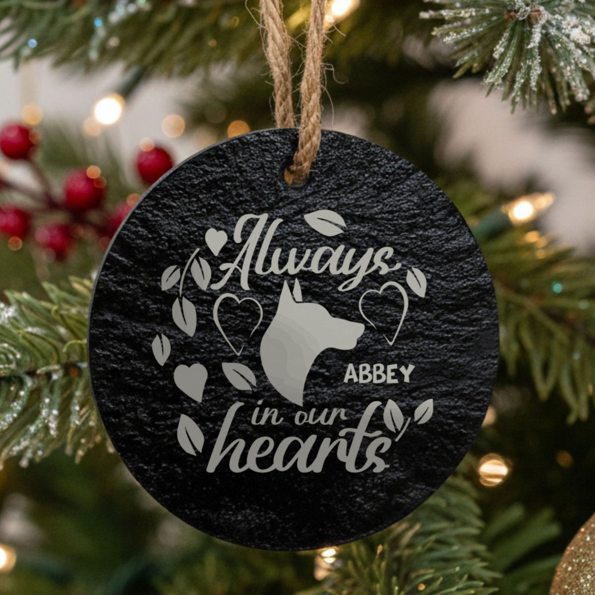 Round slate Christmas ornament engraved Always in Our Hearts with a dog silhouette and the name Abbey, hanging on a tree