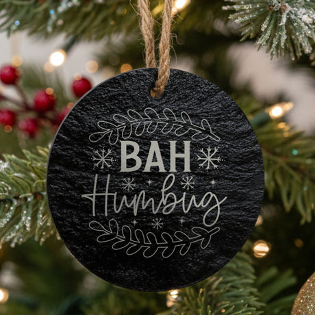 Round black slate Christmas ornament hanging from a jute string on a decorated Christmas tree, laser engraved in white with “Bah Humbug”.