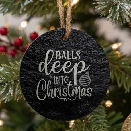 Round black slate Christmas ornament hanging from a jute string on a decorated Christmas tree, laser engraved in white with “Balls Deep Into Christmas”.