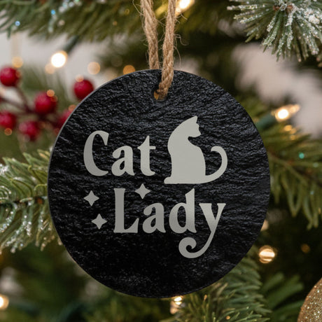 Round black slate Christmas ornament hanging from a jute string on a decorated Christmas tree, laser engraved in white with “Cat Lady”.