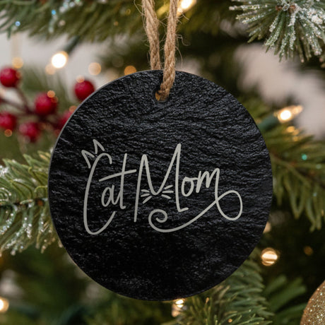 Round black slate Christmas ornament hanging from a jute string on a decorated Christmas tree, laser engraved in white with “Cat Mom Script”.