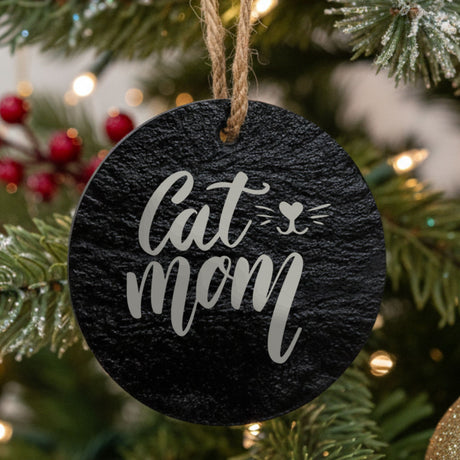 Round black slate Christmas ornament hanging from a jute string on a decorated Christmas tree, laser engraved in white with “Cat Mom Whiskers”.