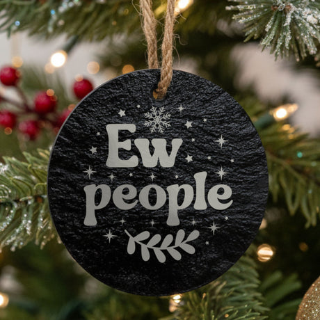 Round black slate Christmas ornament hanging from a jute string on a decorated Christmas tree, laser engraved in white with “Ew People”.