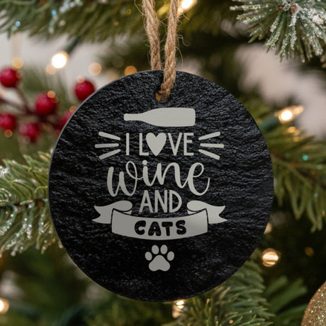Round black slate Christmas ornament hanging from a jute string on a decorated Christmas tree, laser engraved in white with “I Love Wine and Cats”.