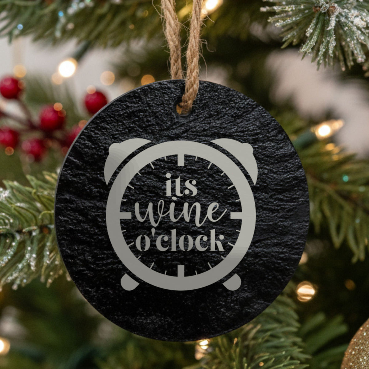 Round black slate Christmas ornament hanging from a jute string on a decorated Christmas tree, laser engraved in white with “It's Wine O'Clock”.