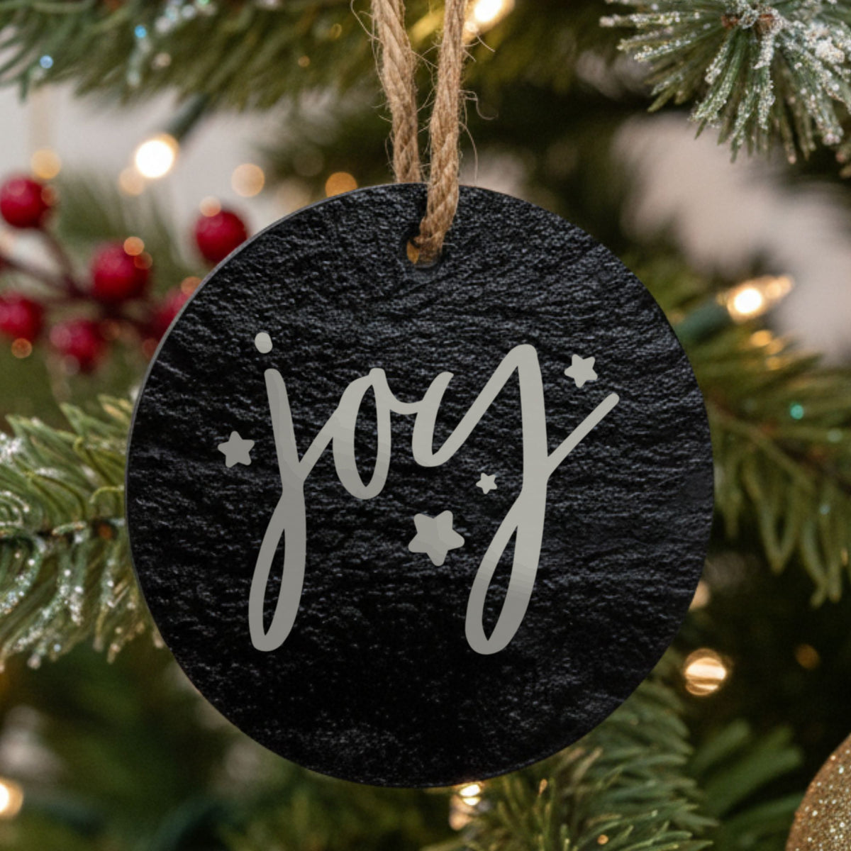 Round black slate Christmas ornament hanging from a jute string on a decorated Christmas tree, laser engraved in white with “Joy”.