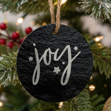 Round black slate Christmas ornament hanging from a jute string on a decorated Christmas tree, laser engraved in white with “Joy”.