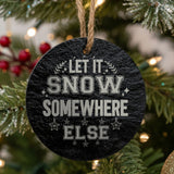 Round black slate Christmas ornament hanging from a jute string on a decorated Christmas tree, laser engraved in white with “Let It Snow Somewhere Else”.