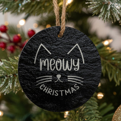 Round black slate Christmas ornament hanging from a jute string on a decorated Christmas tree, laser engraved in white with “Meowy Christmas”.