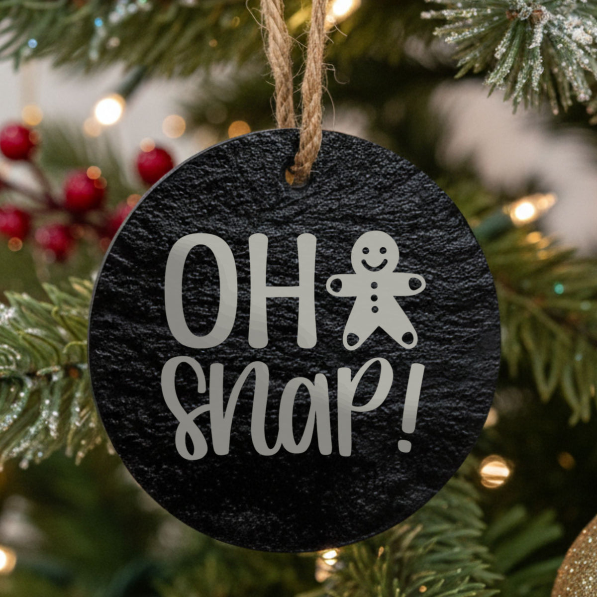 Round black slate Christmas ornament hanging from a jute string on a decorated Christmas tree, laser engraved in white with “Oh Snap”.